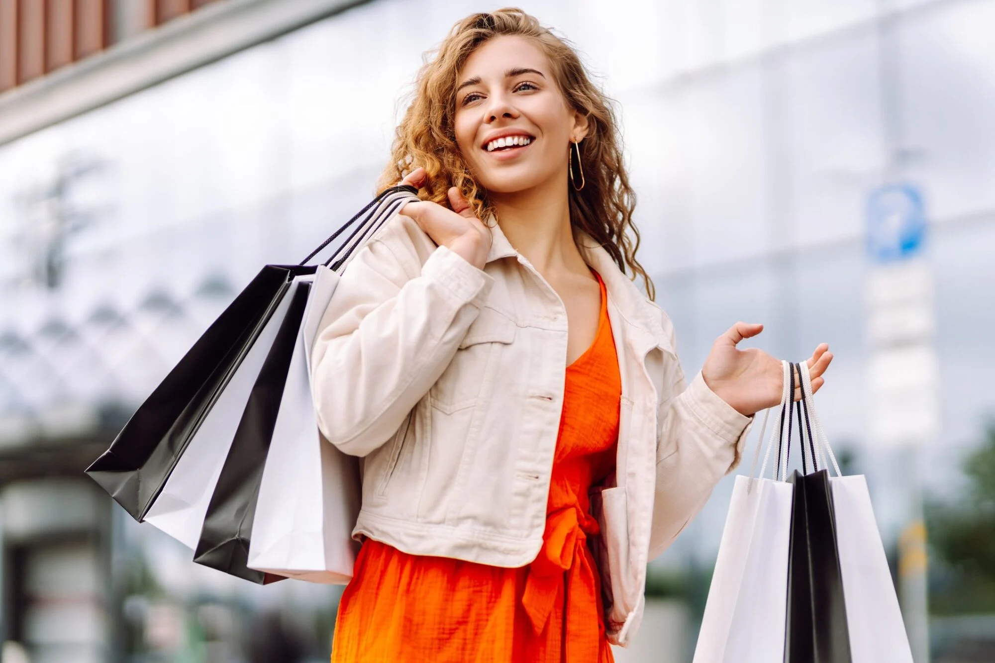 Smiling woman in an orange dress holding shopping bags outdoors.